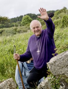 The Bishop of Truro waving. He is in a field with a stone by his side and a person stood behind him. 
