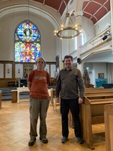 Two people stood in the church with a stained glass window in the background and the halo heater above.