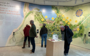 Image shows people inside the Interpretive Centre looking at the displays. The walls have a map on them and there is a wooden display stand in the centre with a glass case on top.