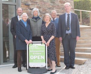 Image shows people stood together outside a stone building. In front of them is a sign which reads 'Interpretive Centre Open'. 