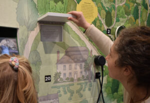 A person on the right looking at something on the wall. The wall is green and there are illustrations on it including a drawing of a house with many windows. Bottom right hand corner shows the back of someone's head.