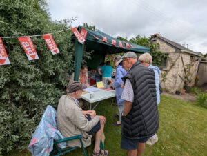 People outside looking at a stall. There are red Christian Aid flags hanging from the bush and a yellow building to the right.