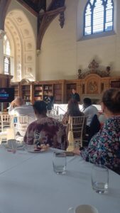 Image shows people in a large room sat at tables looking at a speaker.