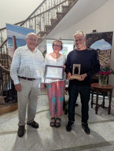 Three people stood holding an award and certificate. There is art and a staircase in the background.