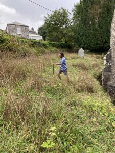 Image shows a grassy churchyard with a person holding a scythe ready to cut the grass. 