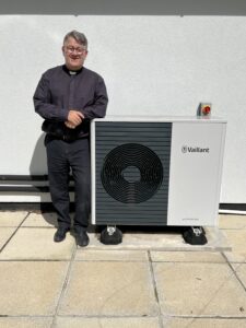 Image shows someone stood next to a heat pump by a white wall and patio.