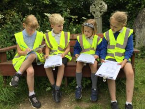 Image shows four children in high-vis vests holding on to clip boards. there is a grave stone and trees behind.