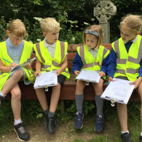 Image shows four children in high-vis vests holding on to clip boards. there is a grave stone and trees behind.