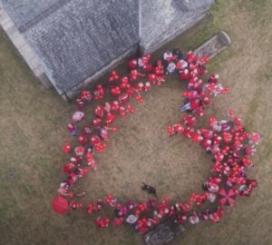 A bird's eye view of people, dressed in red, forming the shape of a heart in the grounds of Mullion Church - it is also a bit misty.