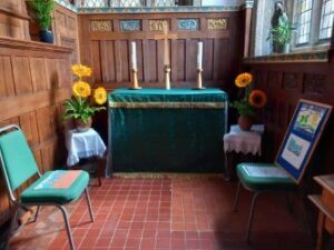 A side chapel with sunflowers either side of a table cloth with green cover and candlesticks on top. the floor is brown in small tiles. there are two green chairs either side.