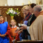 Two children to the left with adults to the right. They are stood in a church with a wooden background.