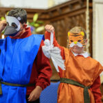 Two children, one in orange with a tiger mask, holding a white prop, and one in blue with a mask.