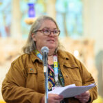 Someone in a light brown jacket reading off paper with a microphone in front of them. The blurred background of the inside of the church and windows.