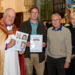 The Bishop of Truro to the left holding a piece of paper with faces on it. He is with with two others - one holding a certificate.