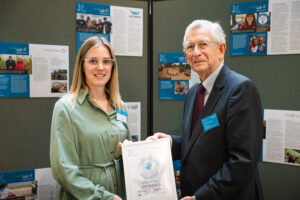 Rebecca St Ledger-Renfree with another and the award in a frame. Background is a display board with posters on it.