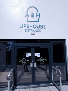 Image shows an entrance to the Lifehouse. There is a white wall with the logo on and underneath are glass doors.