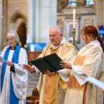 Three people, one is the Bishop of Truro, reading from a book in the cathedral.