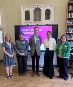 Image shows five people stood in a row on a wooden floor in front of a pink presentation. There is a book shelf to the left.