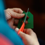 Someone cutting green felt in the shape of holly with red- handle scissors.