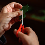 Someone cutting felt into the shape of holly with red handled scissors.