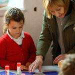 A person in a green coat leaning to help children with crafts. Glue sticks in the foreground. Blue card on the table.