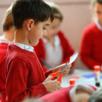 A child cutting paper with red handled scissors. children in the background. A table holds glue sticks.