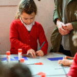 A child is drawing with a person next to them in a green coat. there are children to the sides and a table in the middle holding scissors and glue.