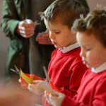 Two children cutting yellow paper with scissors. A person with a green coat stands in the background.