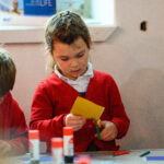 A child is drawing with a person next to them in a green coat. There is a child to the side and a table in the middle holding scissors and glue.