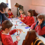 Children around the table cutting with scissors. Three adults helping. A table holds clue, coloured paper, pencils and stars.
