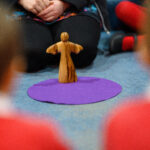 A wooden figure on a purple mat in the centre. A person sits behind it.