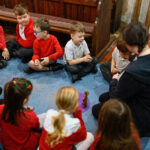 Children sat in a circle on the floor around an adult leading a workshop. Children sat in a circle on the floor around an adult leading a workshop. There is a wooden figure on a purple mat in the centre.