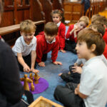 Children sat in a circle on the floor around an adult leading a workshop. There are wooden figures on a purple mat in the centre.