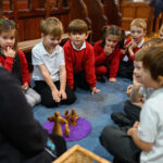 Children sat in a circle on the floor around an adult leading a workshop. There are wooden figures on a purple mat in the centre.