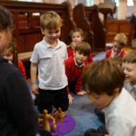 Children sat in a circle on the floor around an adult leading a workshop. There is a wooden figure on a purple mat in the centre.