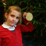 A child holding a handmade Christmas decoration hanging on a tree.