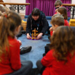 Children sat n a circle with an adult and wooden figures on a purple mat.