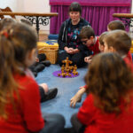 Children sat n a circle with an adult and wooden figures on a purple mat.