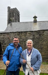 Two people stood in front of a brick wall with roof and spire. The people are holding sticks.