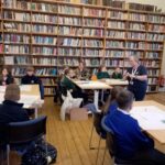 Children sat at desks with a large and full bookshelf in the background.
