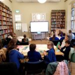 Children sat at desks with a large and full bookshelf and a tv screen in the background.