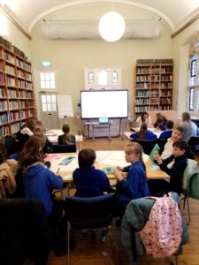Children sat at desks with a large and full bookshelf and a tv screen in the background.