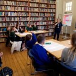 Children sat at desks with a large and full bookshelf in the background.