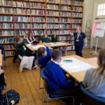Children sat at desks with a large and full bookshelf in the background.