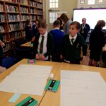 Children looking at paper on a table with a large bookshelf to the right.