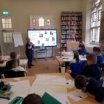 Children sat at desks with a large bookshelf, Clare Green and a TV screen in the background.