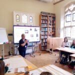 Children sat at desks with a large bookshelf, Clare Green and a TV screen in the background.