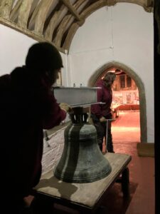 A silhouette of a person in the front left carrying a bell towards an archway. The arched roof is made of wood and the walls are white. the floor is reddish, brown. 