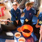 Children watching a workshop demonstration before they give it a go. There is an orange bucket in the centre.