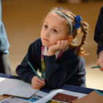 A small child with their head in their hands looking up. They are holding a pencil ready to write.